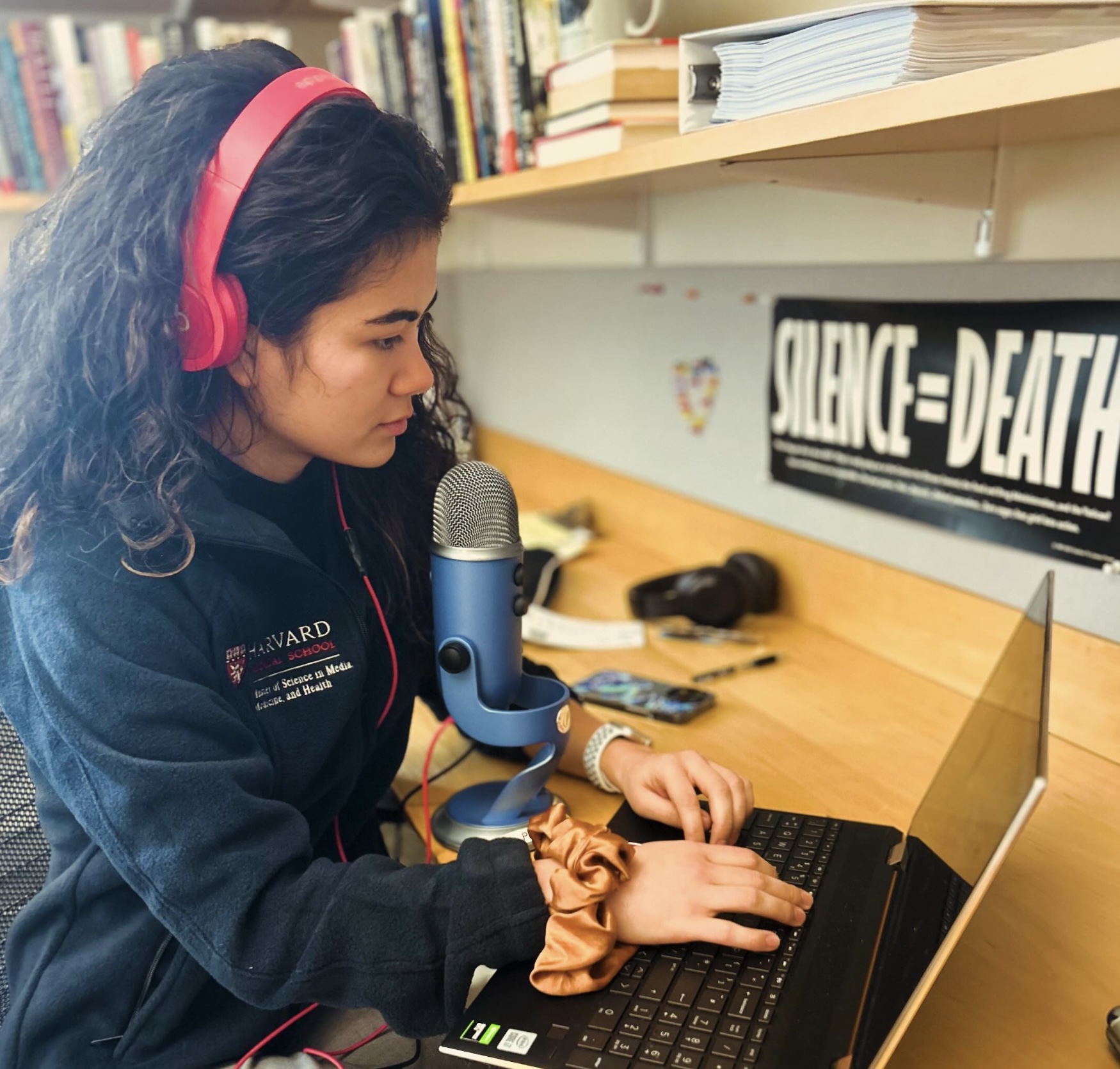 Nicola Sharp sits in an office with her laptop wearing headphones in front of a microphone. A sign on the wall reads silence equals death.