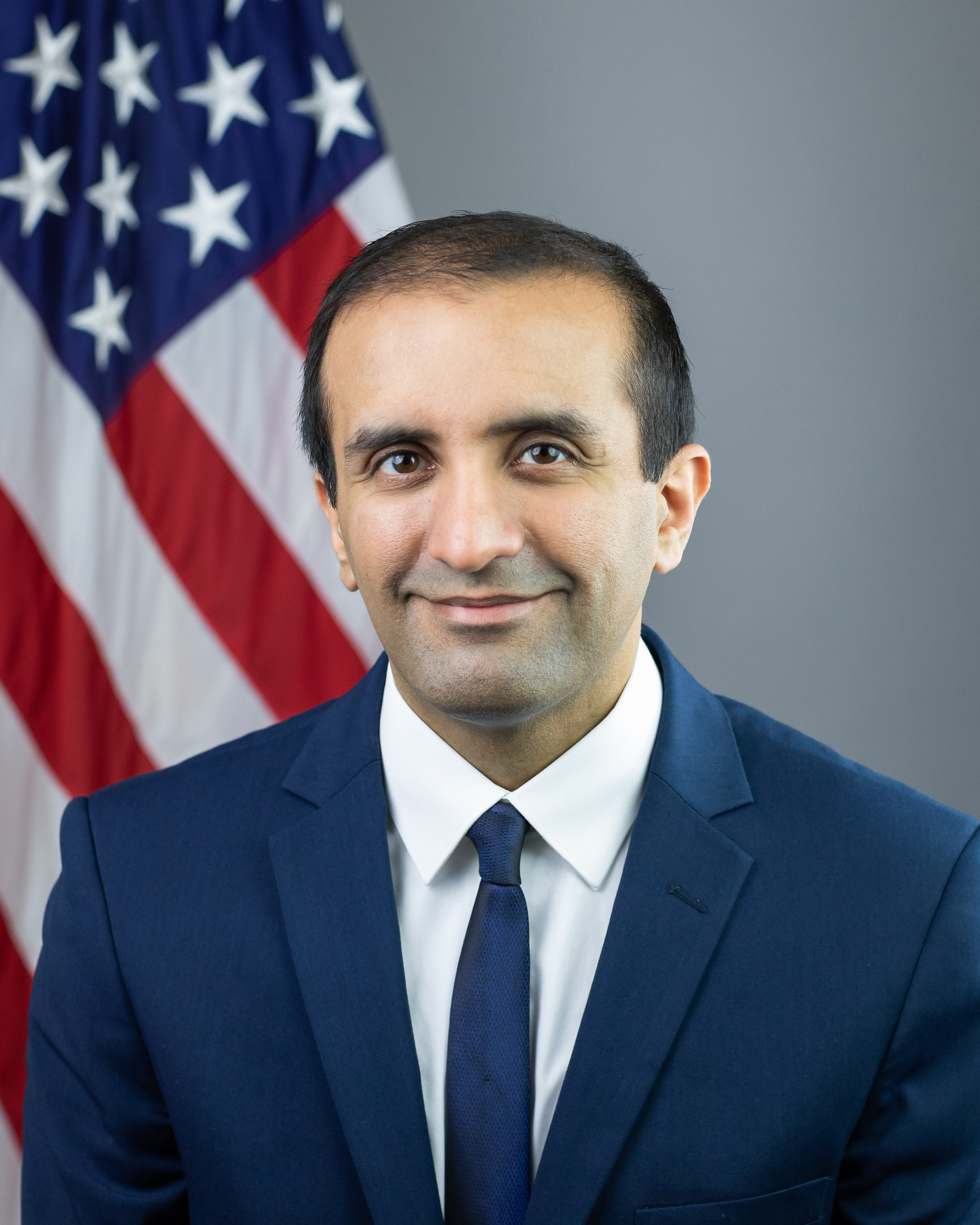 Dr. Raj Panjabi, blue suit and tie, headshot in front of American Flag
