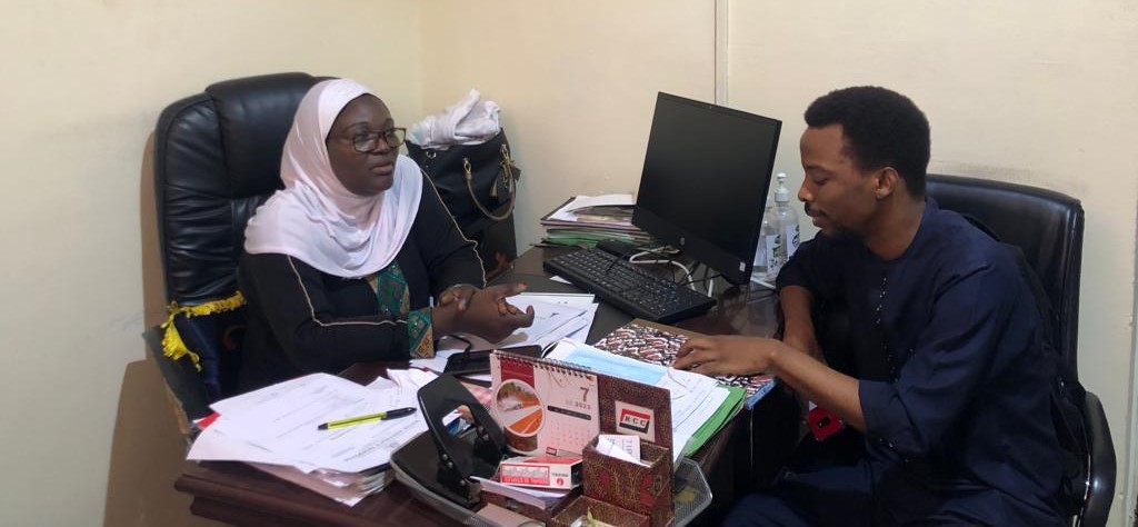 Olamide, seated right. interviews a Ghanan hijabi woman. She is seated behind a desk cluttered with papers. 