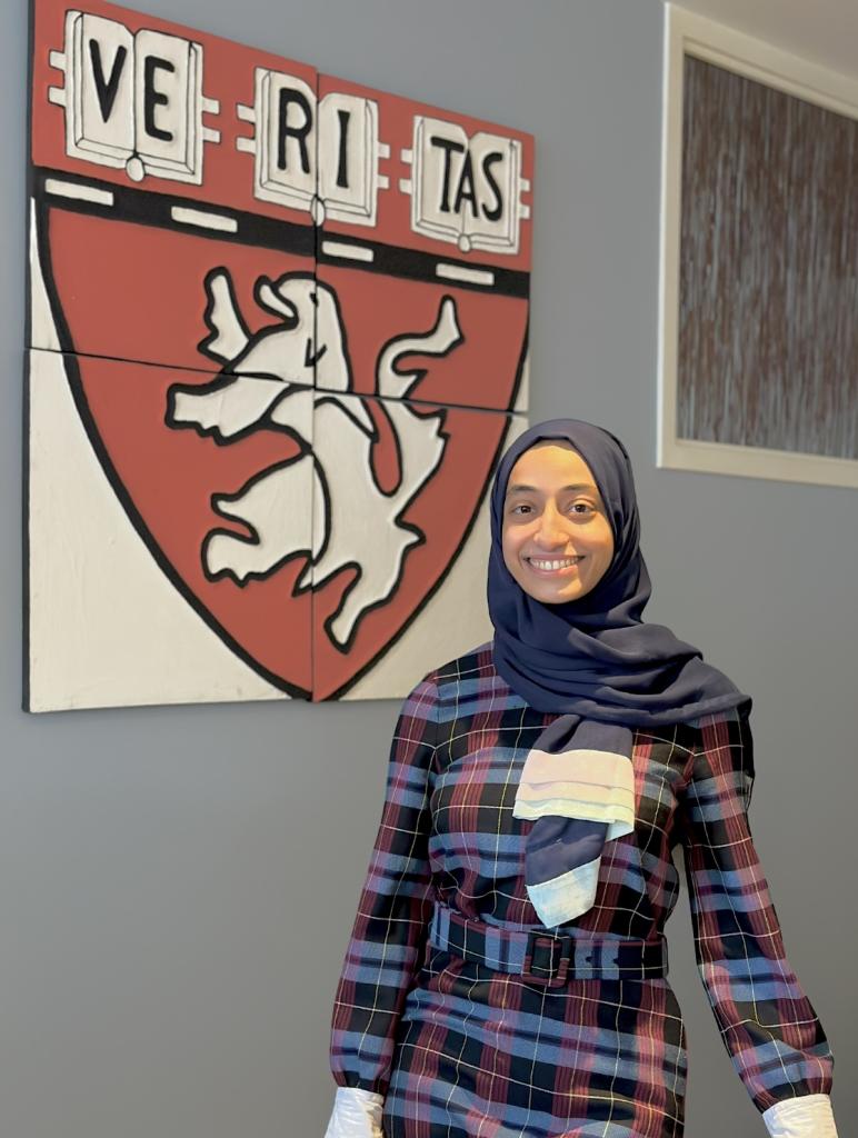 Razan Baabdullah stands smiling in the front lobby of the Department of Global Health and Social Medicine, wearing a plaid shirt and a blue hijab. Behind her is a artistic depiction of the Harvard Medical School shield. 