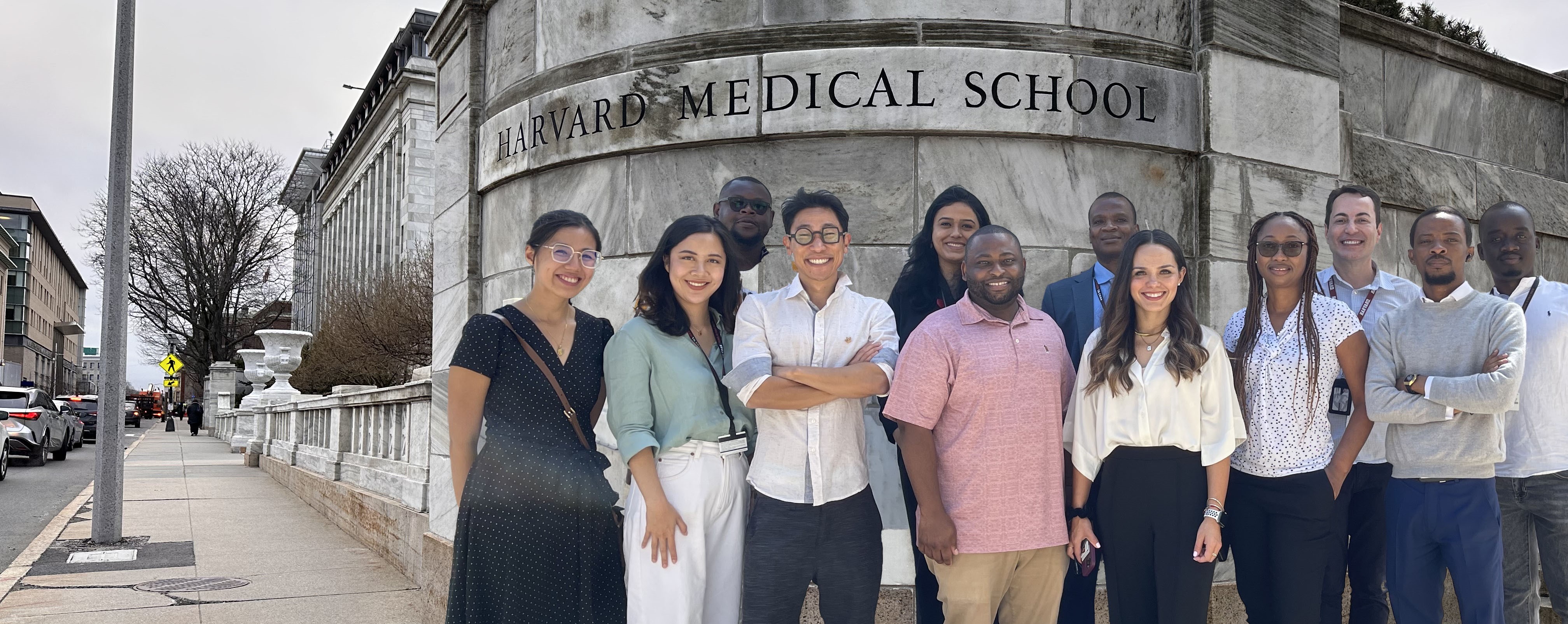 13 students of various ages, genders and nationalities of the GHD Class of 2024 standing outside in front of Harvard Medical School sign etched into marble. 