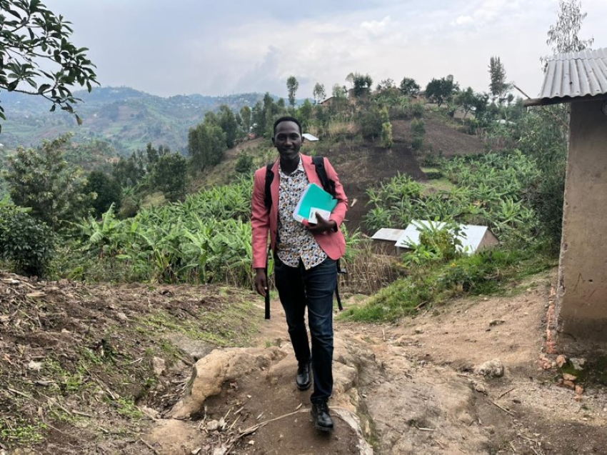 Alain walking over a dirt hill, the green brush hills of Rwanda behind him. He holds a binder filled with research 