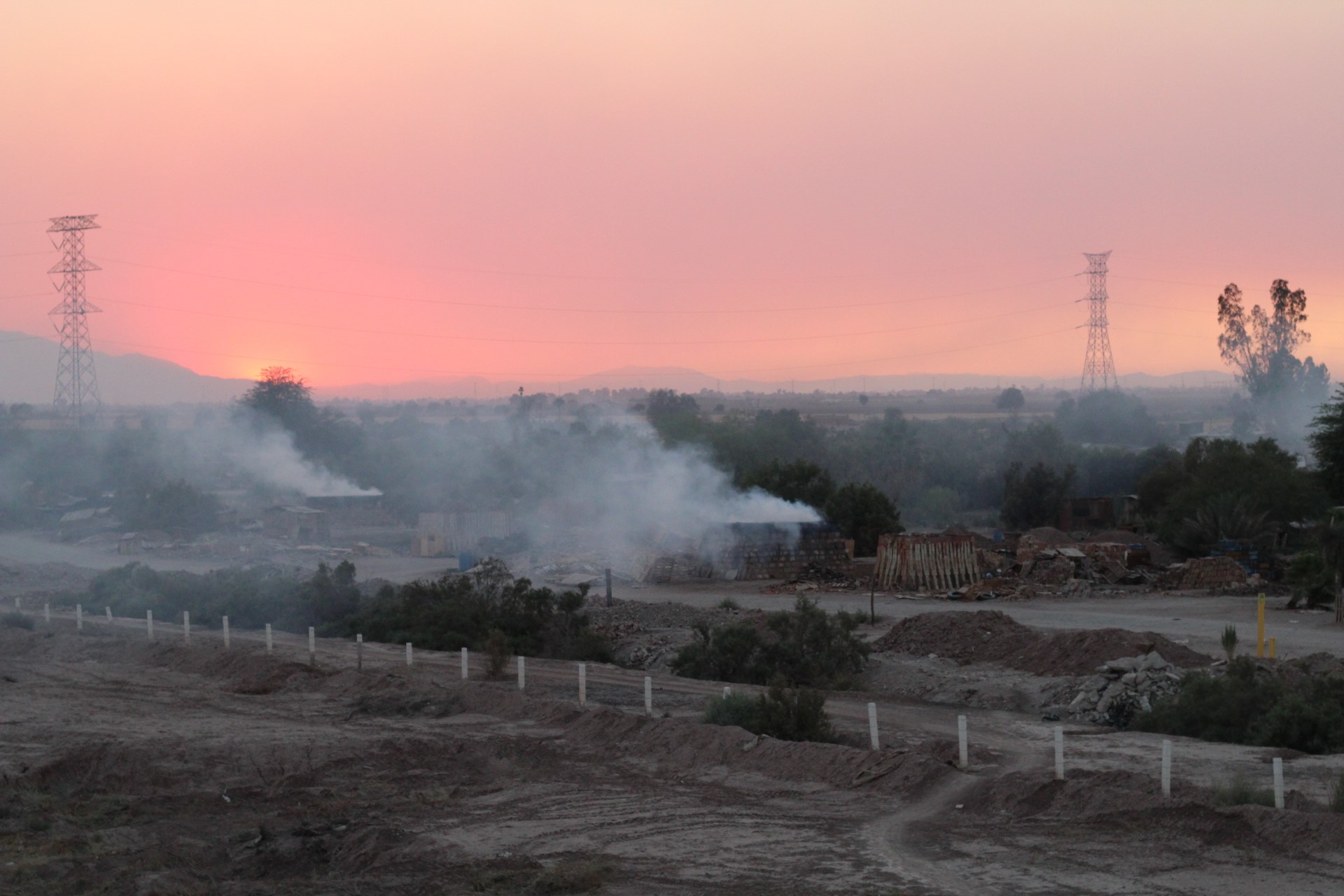 A sun sets behinds the hills of La Ladrillera Mexico. Smoke comes out of a brick kiln, covering the sunset. The foreground is a pile of dirt
