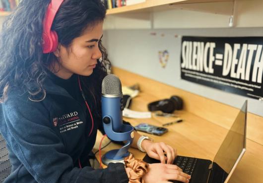 Nicola Sharp sits in an office with her laptop wearing headphones in front of a microphone. A sign on the wall reads silence equals death.