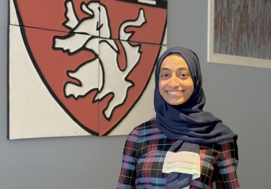 Razan Baabdullah stands smiling in the front lobby of the Department of Global Health and Social Medicine, wearing a plaid shirt and a blue hijab. Behind her is a artistic depiction of the Harvard Medical School shield. 