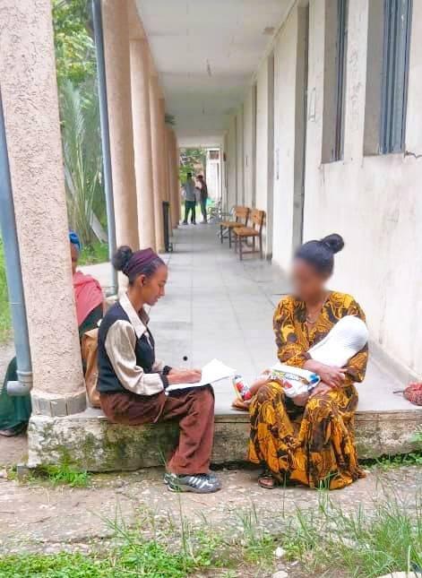 Two women sit on a porch. One has a notebook. The other has a baby in her arms